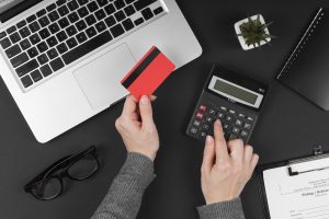 A person is holding a red credit card in one hand while using a calculator with the other, surrounded by a laptop, black-framed glasses, a small potted plant, a spiral notebook, and a clipboard with a delivery form on a dark background. The scene suggests financial planning, online shopping, or budgeting activities.