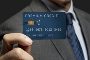 Close-up of a businessman in a suit holding a blue premium credit card with contactless payment symbol, displaying the name William Jones and card details.