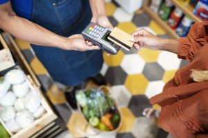 A customer in a rust-colored blouse pays for fresh groceries at a small shop using a contactless method with a gold-colored credit card. A shopkeeper in a denim apron holds the card reader, highlighting the everyday use of credit cards for in-store purchases.