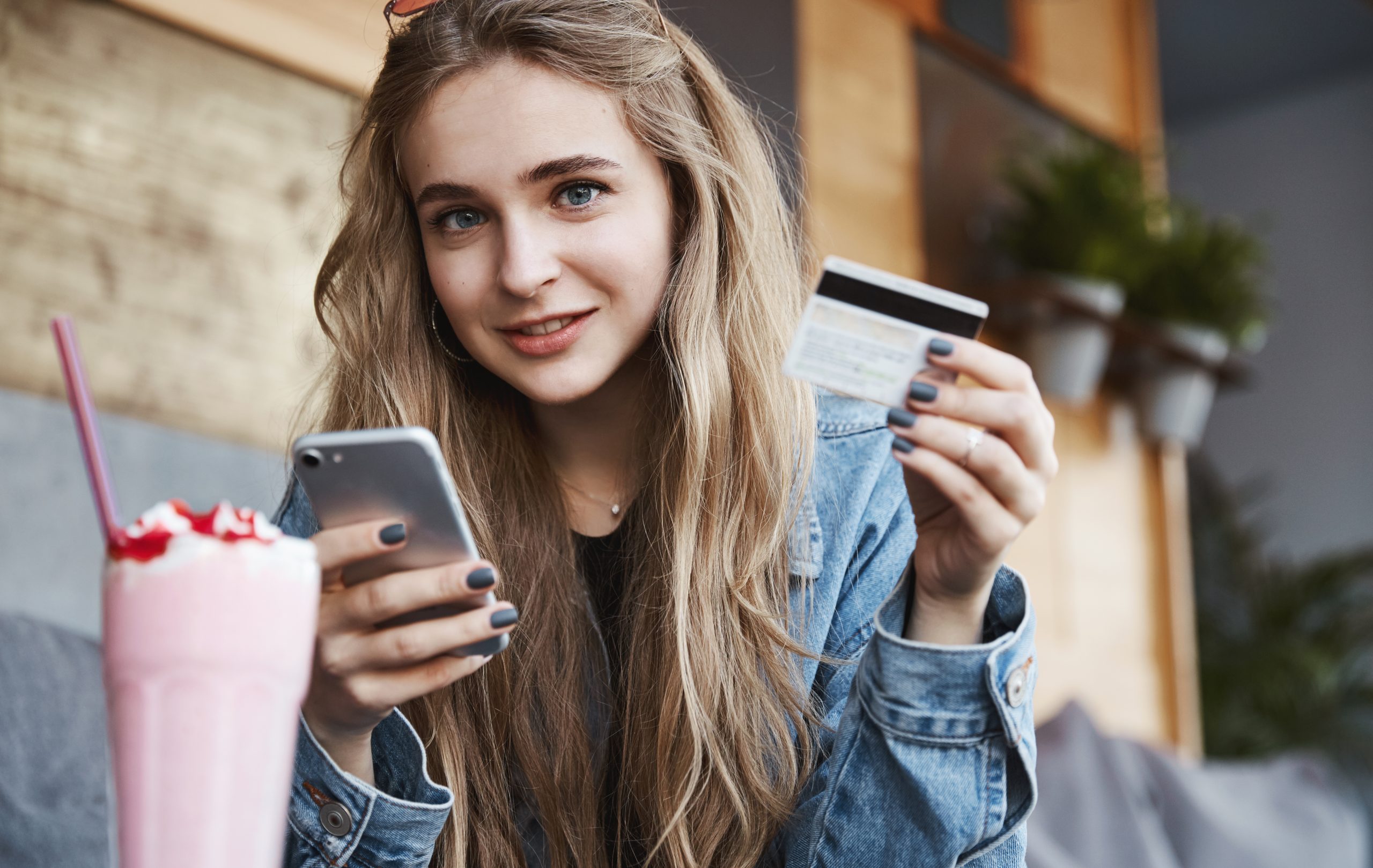 A young woman with long blonde hair, wearing a denim jacket, is sitting at a café holding a credit card in one hand and a smartphone in the other. She has a friendly smile and appears to be making an online payment or purchase. A pink milkshake with whipped cream and a straw sits on the table in front of her. The background features a cozy café setting with wooden decor and potted plants, creating a warm and inviting atmosphere.