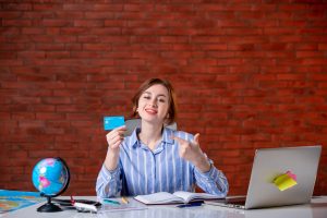 A young woman in a striped blue shirt is sitting at a desk, smiling and pointing at a blue credit card she is holding. The desk is covered with travel-related items, including a globe, a map, an open notebook, a laptop with sticky notes attached, and some stationery. The red brick wall in the background adds a cozy and modern touch to the scene, suggesting themes of travel planning, financial management, or online booking.