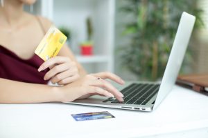 A woman wearing a burgundy dress is using a laptop while holding a yellow credit card in one hand and typing with the other. A second credit card is placed on the white desk beside her. The background features a bright, modern indoor setting with green plants and decorative elements, suggesting online shopping, digital payments, or financial transactions.