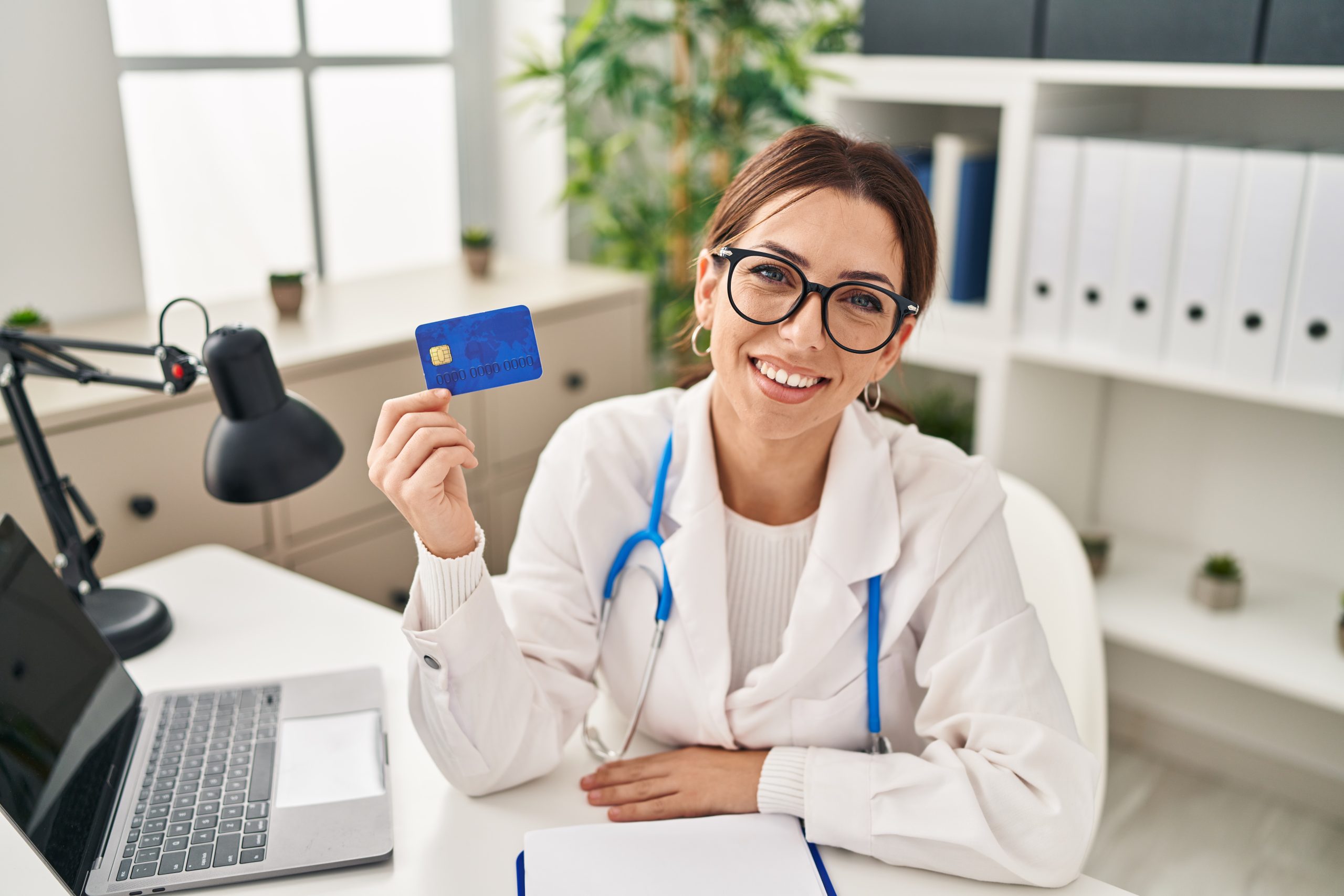 A young female doctor with brown hair, wearing glasses and a white coat with a stethoscope around her neck, is sitting at a desk in a medical office. She is smiling and holding a blue credit card, suggesting themes of medical payments, health insurance, or financial transactions in healthcare. The background features a well-organized office with shelves, binders, plants, and a laptop on the desk, creating a professional and welcoming atmosphere.