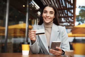 A young woman with long dark hair, wearing a beige turtleneck and a light gray coat, is sitting at a table in an outdoor coffee. She is smiling as she holds a credit card in one hand and a smartphone in the other, suggesting that she is making a payment or an online purchase. On the table is a takeaway coffee cup and the background features a modern coffee setting with wooden details and warm lighting.