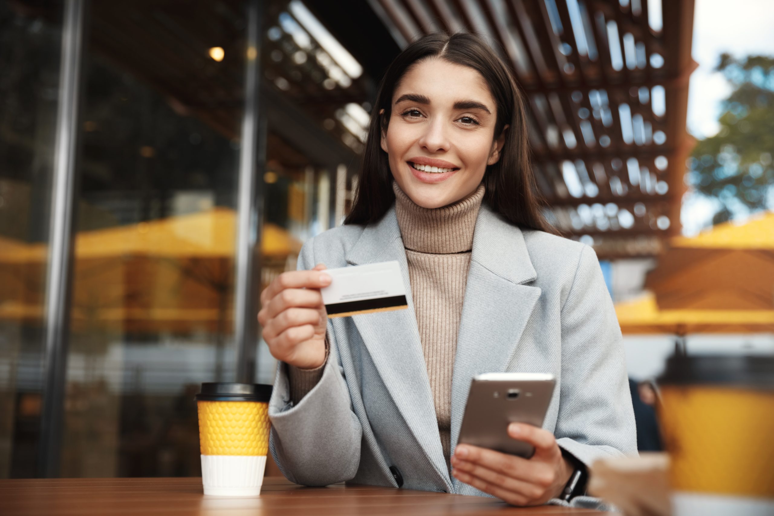 A young woman with long dark hair, wearing a beige turtleneck and a light gray coat, is sitting at a table in an outdoor coffee. She is smiling as she holds a credit card in one hand and a smartphone in the other, suggesting that she is making a payment or an online purchase. On the table is a takeaway coffee cup and the background features a modern coffee setting with wooden details and warm lighting.