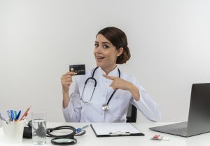 A young female doctor in a medical coat holding a credit card and pointing at it while sitting at a desk with medical tools and a laptop, smiling and looking impressed.