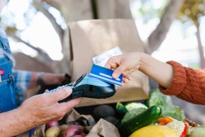 A person making a payment with a credit card at a market stand, while purchasing fresh produce, with a payment terminal in the other person's hand.