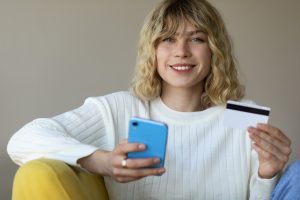 Smiling young woman sitting on a yellow chair, holding a smartphone in one hand and a credit card in the other, making an online purchase.