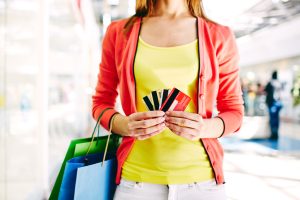 A woman standing in a shopping mall, holding multiple credit cards in both hands and carrying colourful shopping bags, dressed in a yellow top and coral cardigan.