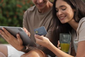 A young couple sits outdoors, smiling while shopping online using a tablet and a modern chip-enabled card. The woman holds a glass of juice and a credit card, while the man navigates on the device—illustrating the digital habits and financial engagement of Credit Cards and Generation Z.