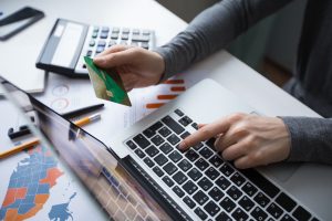 A person sits at a desk using a laptop for online financial transactions, holding a green credit card in one hand while typing with the other. Surrounding documents and charts indicate economic analysis, visually representing the impact of variable interest rates on credit cards and personal budgeting.