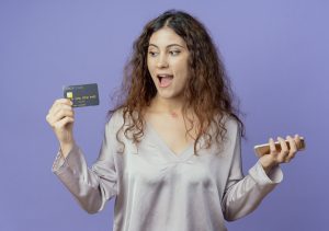 A young woman with curly brown hair, wearing a silky light gray blouse, is holding a black credit card in one hand and a smartphone in the other. She has an excited and surprised expression on her face as she looks at the card. The background is a solid purple color, creating a modern and vibrant setting that suggests online shopping, financial success, or digital transactions.