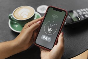 A person holds a smartphone displaying a "Pay" button and a shopping cart icon, ready to complete a transaction using NFC technology. A contactless card reader and a cup of coffee are visible on the table, illustrating the convenience of digital payments in everyday purchases.
