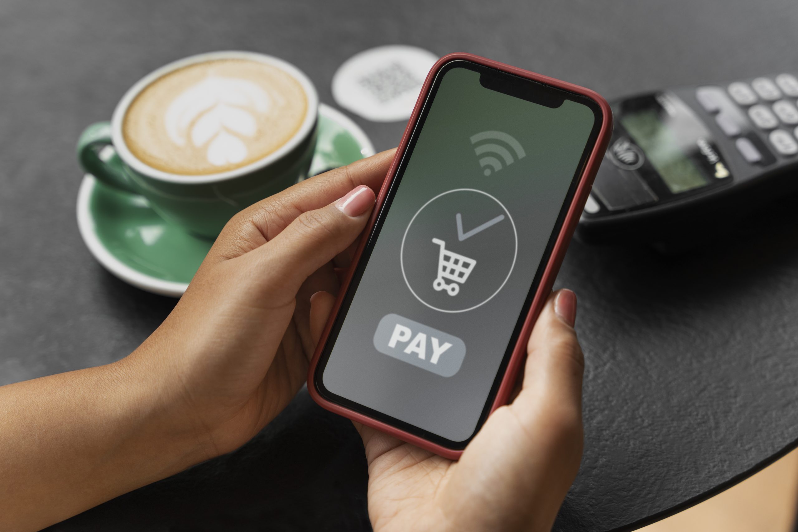 A person holds a smartphone displaying a "Pay" button and a shopping cart icon, ready to complete a transaction using NFC technology. A contactless card reader and a cup of coffee are visible on the table, illustrating the convenience of digital payments in everyday purchases.