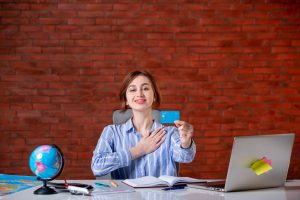 A smiling young woman sits at a desk with a laptop, notebook, and world globe, proudly holding up a blue credit card. Surrounded by educational materials, the scene highlights the use of credit cards to invest in education and professional courses, empowering learners to access knowledge and career opportunities.