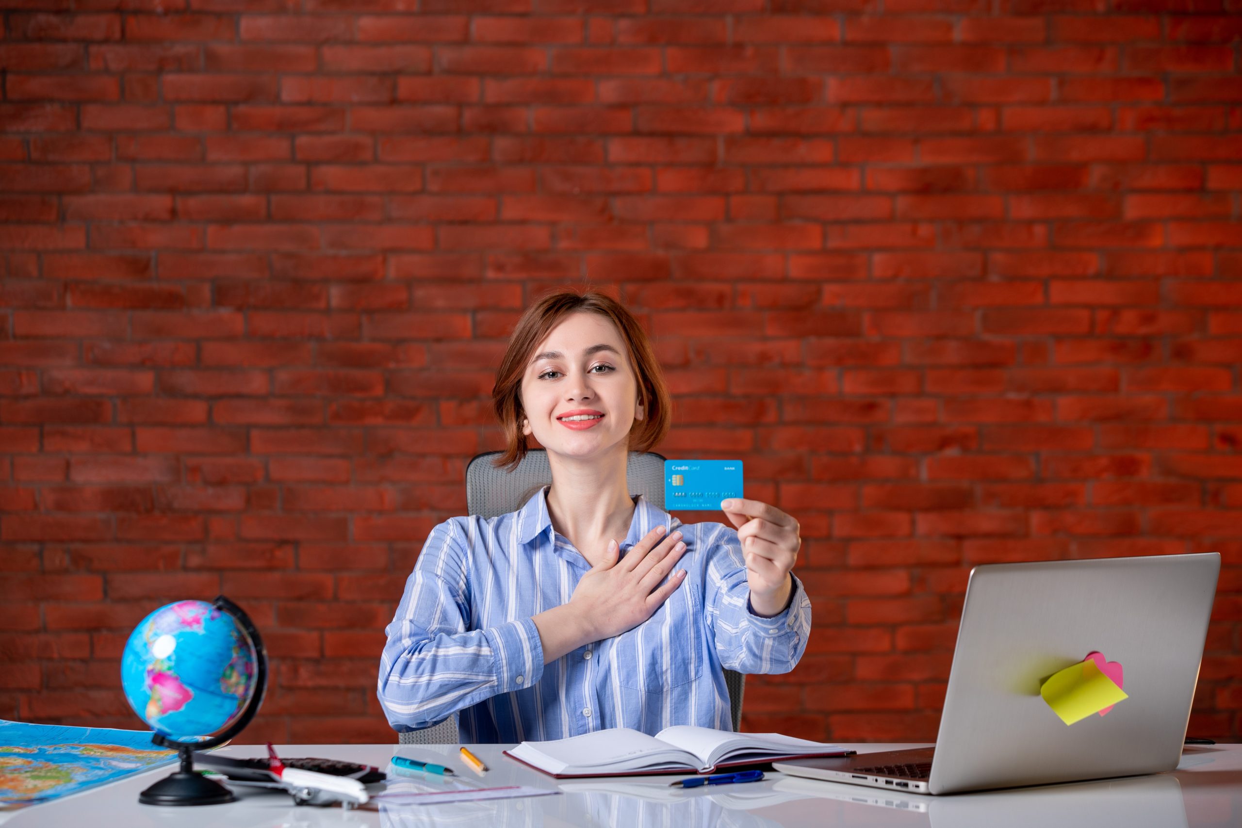 A smiling young woman sits at a desk with a laptop, notebook, and world globe, proudly holding up a blue credit card. Surrounded by educational materials, the scene highlights the use of credit cards to invest in education and professional courses, empowering learners to access knowledge and career opportunities.