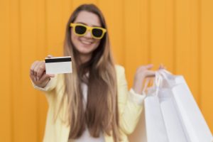 A smiling woman wearing yellow sunglasses and a light yellow blazer stands in front of a vibrant orange background. She holds multiple white shopping bags in one hand and extends a blank credit card toward the camera with the other, emphasizing the use of credit cards for shopping.