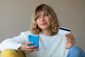 A young woman sitting casually while holding a smartphone and a credit card, reflecting the digital-first consumption behavior of millennials in modern shopping habits.