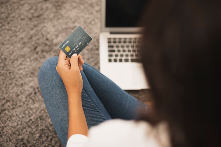A woman sitting cross-legged on the floor in front of a laptop, holding a renewal credit card in her hand while preparing for an online payment.