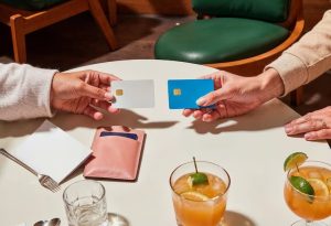 Two people seated at a round table exchanging credit cards over drinks, with a pink wallet, cutlery, and glasses visible in a relaxed, indoor setting.
