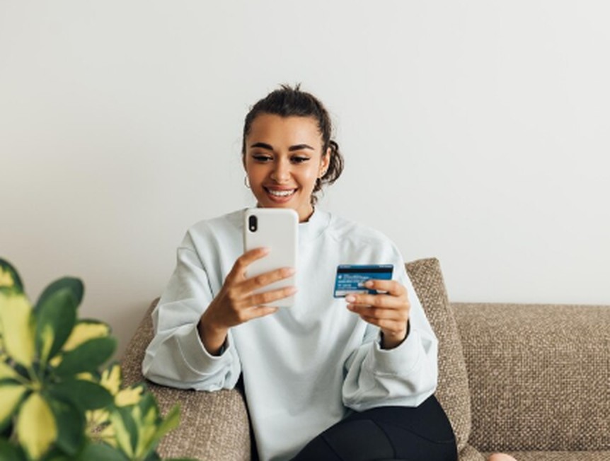A smiling woman sits on a couch holding a smartphone and a debit card, representing the ease of online shopping and the growing popularity of Cashback Debit Cards for everyday purchases.