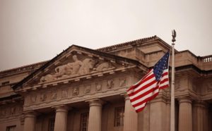 The American flag waves in front of a classical government building with columns and sculptures, symbolizing institutions like the Federal Reserve and their role in U.S. economic policy.