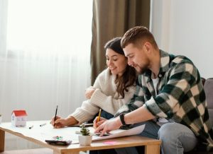A young couple sits at a coffee table reviewing documents and using a calculator, illustrating teamwork and financial planning for shared goals and household budgeting.