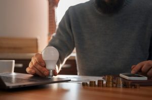 A person sitting at a desk holding a light bulb near a laptop, with stacks of coins and a smartphone nearby, symbolizing ideas and strategies for generating passive income.