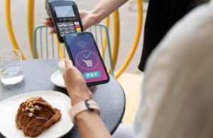 A person at an outdoor café using a smartphone for contactless digital payments, with a pastry and drink on the table.