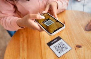 A person uses a smartphone to scan a printed QR code on a wooden table, demonstrating the ease and convenience of QR Code Payments in a real-world setting.