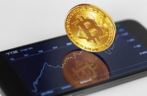 A golden Bitcoin symbol above a smartphone screen displaying a financial chart, representing the rise and volatility of cryptocurrencies.