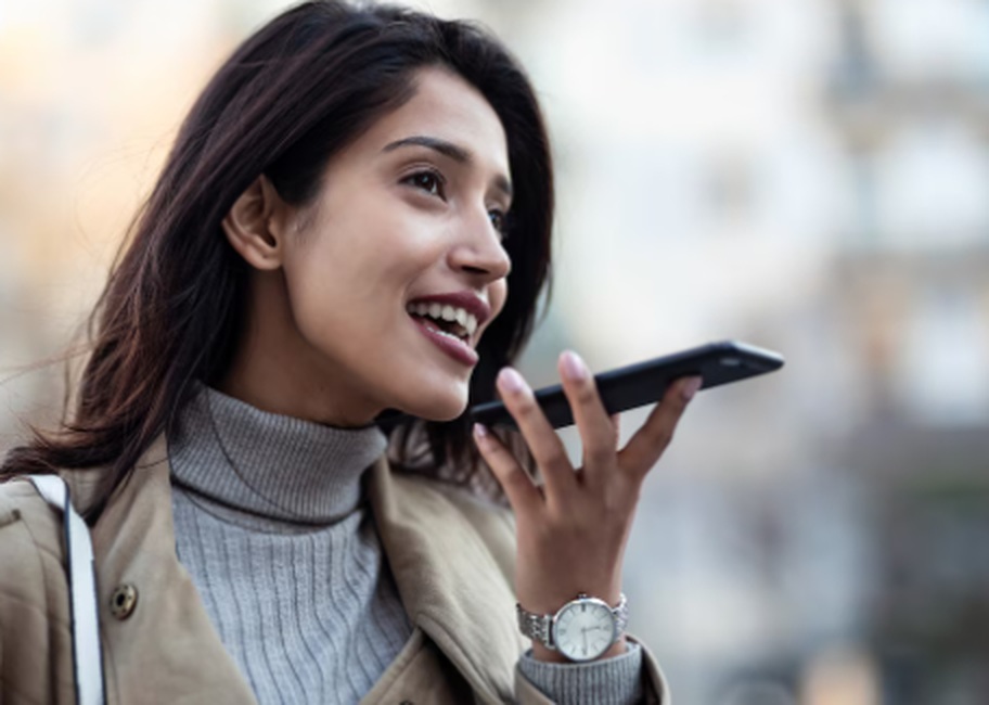 A young woman outdoors smiling while speaking into her smartphone, illustrating the convenience and accessibility of voice payments technology.